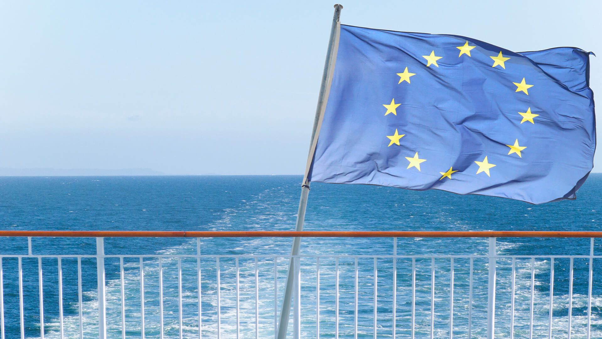 A European Union flag flies from a ship’s railing with the open sea in the background.
